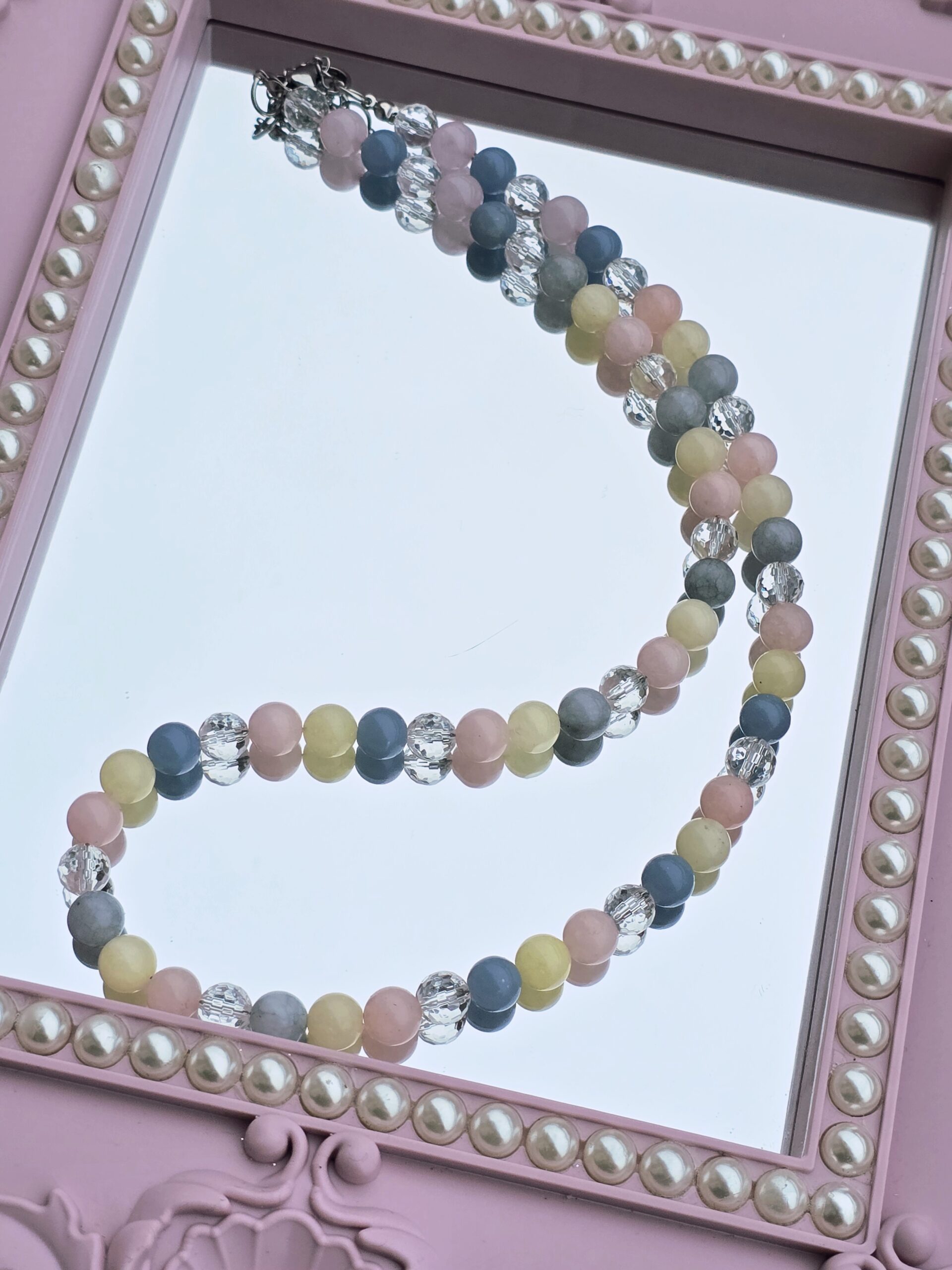 Picture shows a handmade beaded necklace in pastel colors sitting over a mirror with pink frame