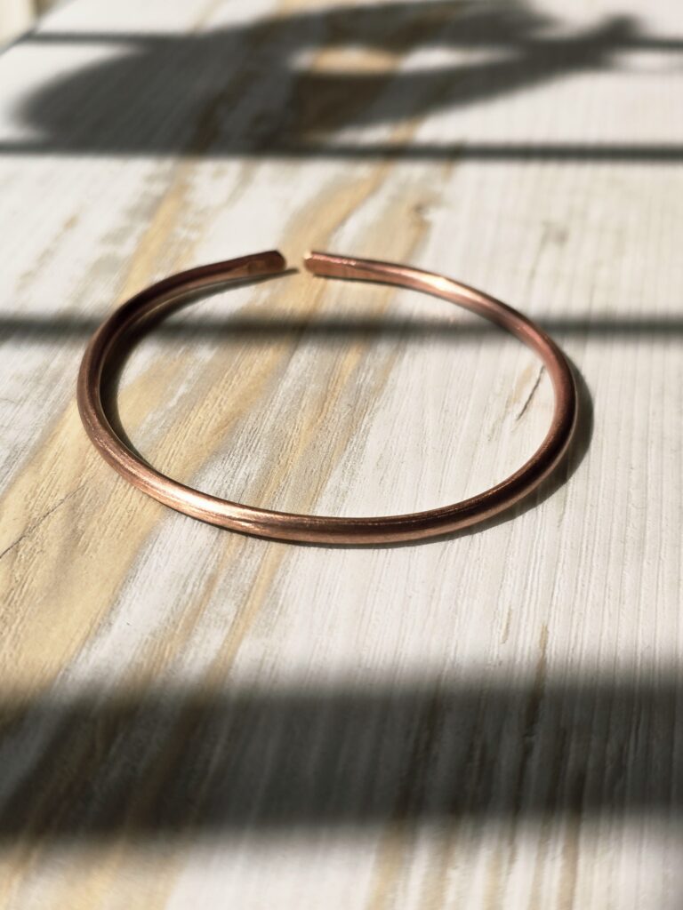 A close‑up of a round copper bangle resting on a sunlit wooden surface. The soft shadows and natural grain emphasize the bracelet’s smooth finish and warm reddish tone