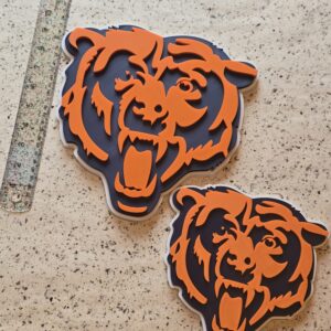 A pair of 3D‑printed bear head emblems in bold orange and navy blue, displayed on a speckled surface with a ruler for scale. Each piece showcases layered precision and clean edges, perfect for sports fans and custom décor enthusiasts.