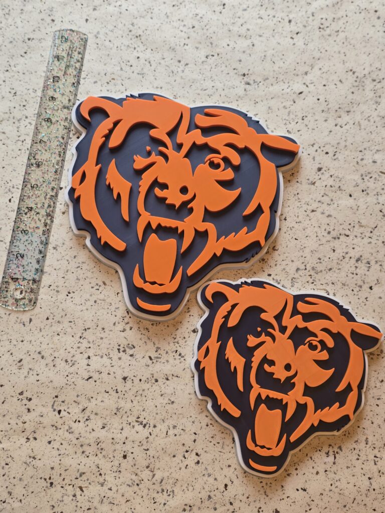 A pair of 3D‑printed bear head emblems in bold orange and navy blue, displayed on a speckled surface with a ruler for scale. Each piece showcases layered precision and clean edges, perfect for sports fans and custom décor enthusiasts.