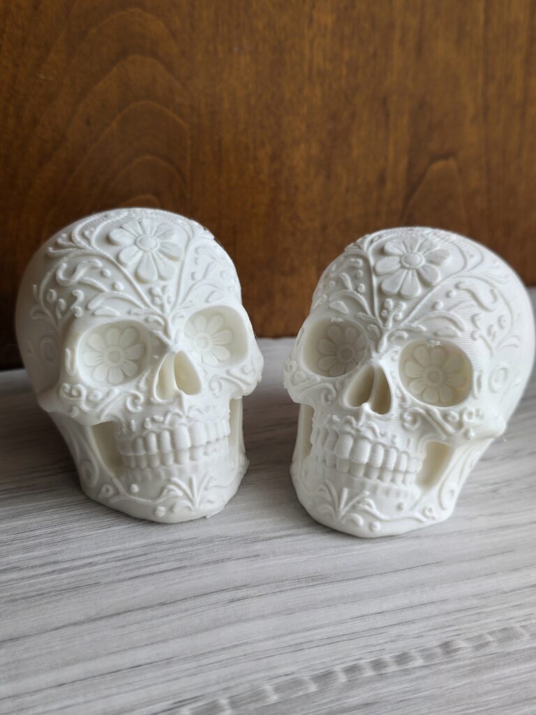 A pair of 3D‑printed sugar skulls in smooth white finish, featuring intricate floral and swirl engravings. The detailed craftsmanship highlights the Day of the Dead‑inspired design, perfect for collectors or festive décor.