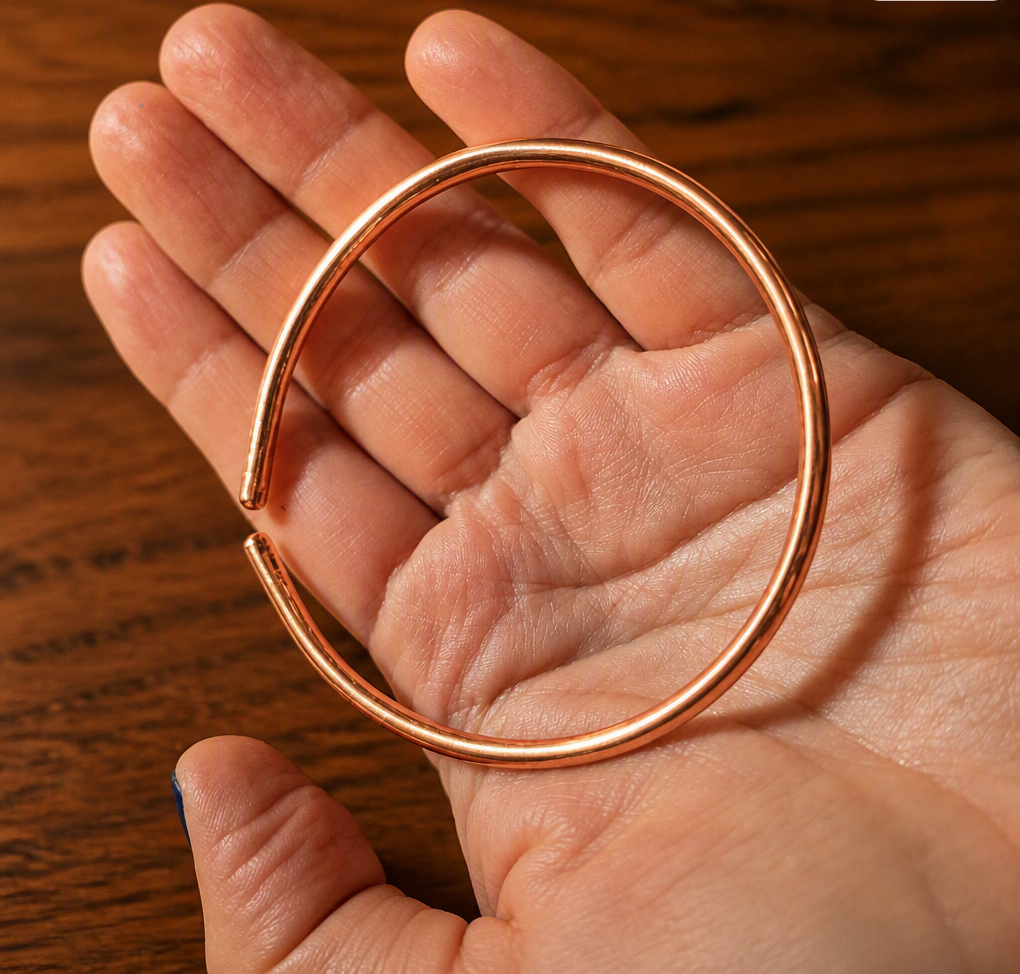 A detailed photo of a polished copper bangle held in the palm, highlighting its smooth curvature and rich metallic sheen. The warm wood background enhances the natural beauty of copper, ideal for artisan jewelry listings and handmade accessories.