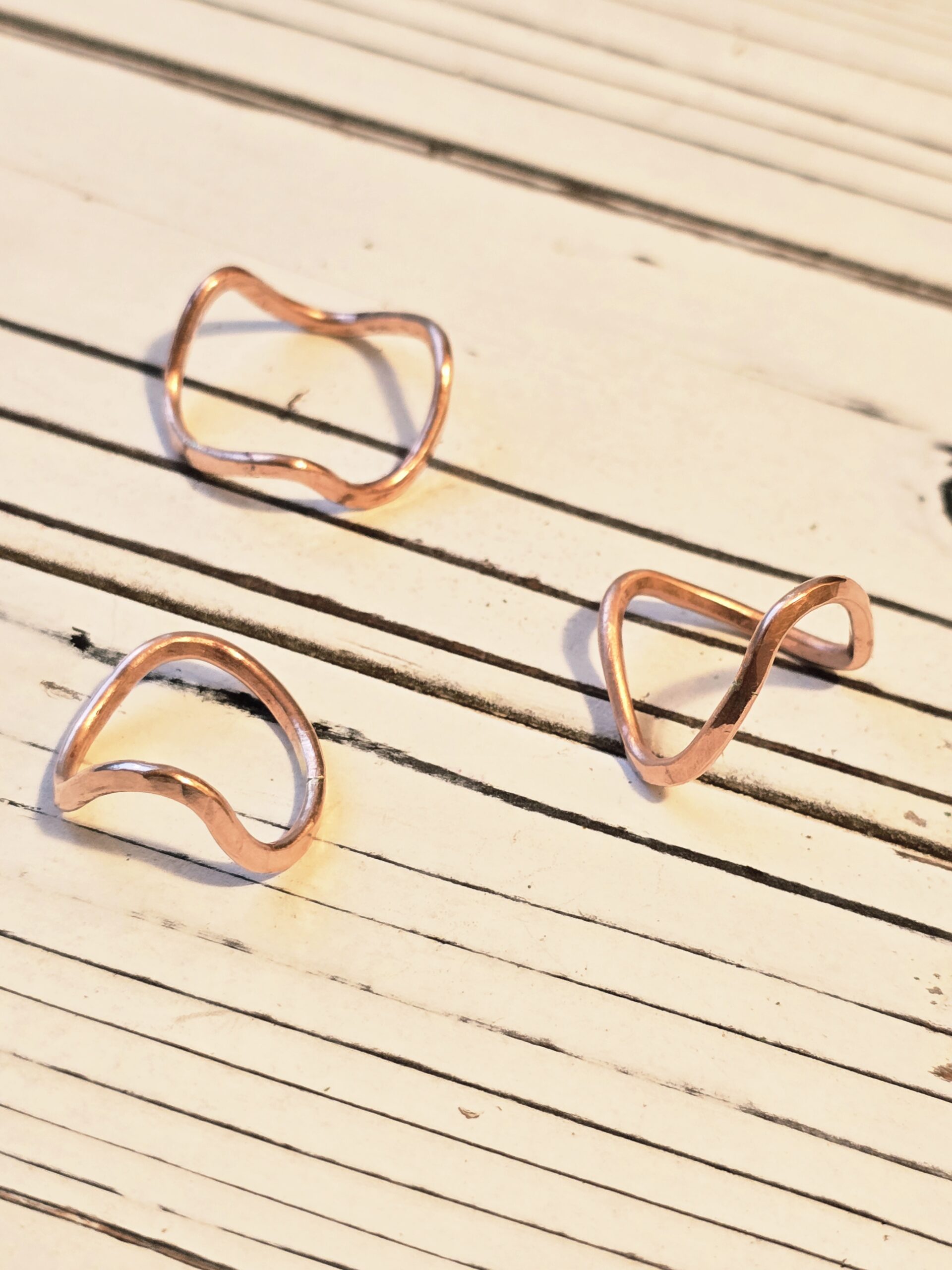 Three hand‑forged copper rings displayed on a rustic wooden background. Each band features a unique wave shape and polished finish, emphasizing individuality and handcrafted artistry.
